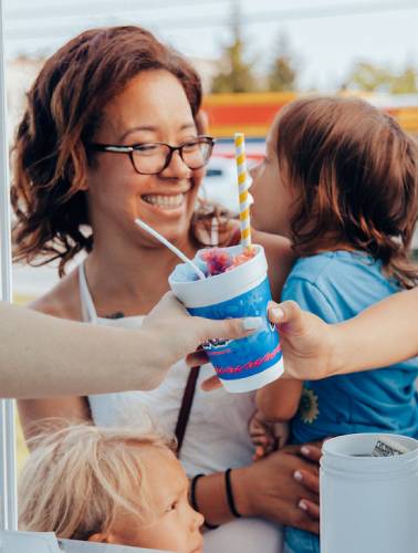 Mom holding child behind hand passing shaved ice